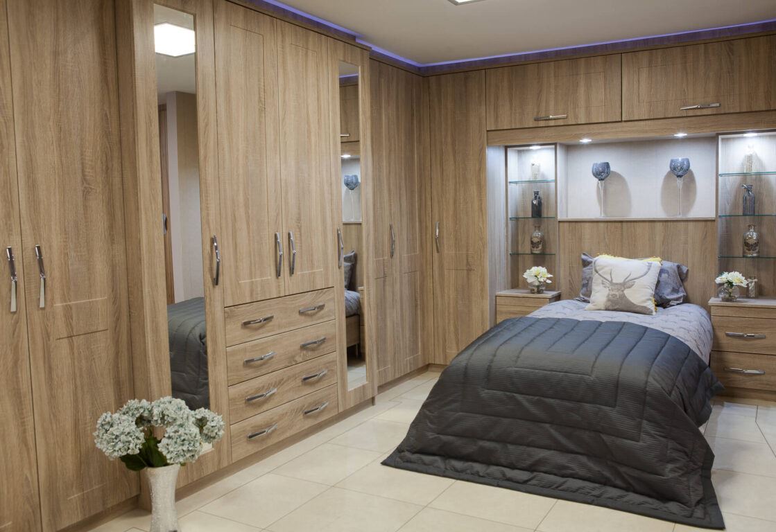 Oak wood effect bedroom furniture, floor to ceilin fitted furniture, overhead pull out storage, glass shelving, and matching bedromm tables for a unified look.
