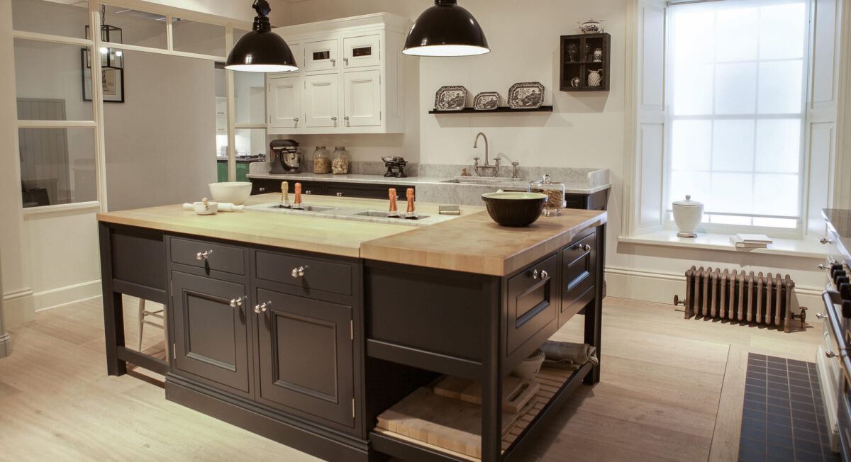 A black kitchen island with a wooden worktop is lit up by two black pendant lights hanging above. There is a ceiling white shaker cabinet and grey quartz worktops