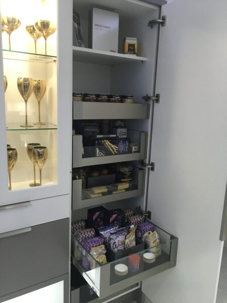 A Cavendish designed Kitchen. Light grey full cabinet open to show pull out larder storage with drawers. Next to it is a cabinet with glass panelling showing open shelving with glassware.