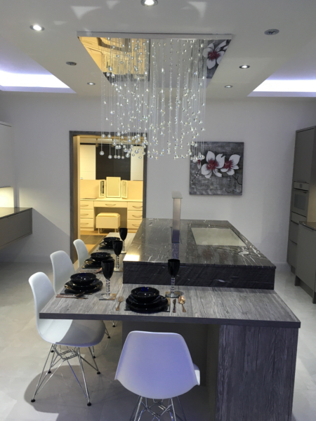 A Cavendish designed Kitchen. Bar stools at kitchen kitchen island area with dark wood surfaces and black cabinets. Island has two levels, a champagne bath and chandelier pendant lighting above.