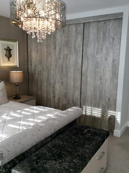A Cavendish designed Bedroom with custom wardrobe in ash wood effect. Chandelier lighting hangs over a crushed velvet bed and chest of drawers set.