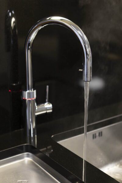 Close-up of a silver curved swan neck hot tap and integrated sink, against a backdrop of a black kitchen splashback and dark marble kitchen work surfaces