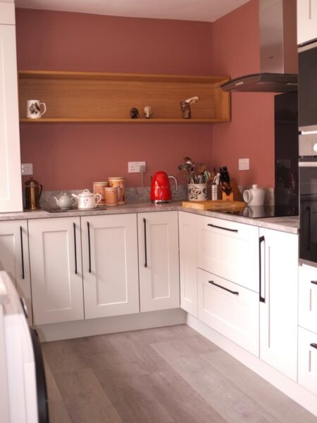 Cavendish kitchen design with rose pink cabinetry and black fixtures. The deep red walls contrast nicely for a fun, kitschy vibe. Open shelves showcase crockery and glassware, adding to the charm.